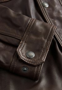 Manschette einer braunen Lederjacke mit einem dekorativen Metall-Druckknopf, Kontrastnähte und einer strukturierten Oberfläche, sichtbar gefaltet mit Details.