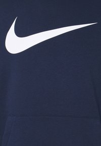 Logo Nike swoosh blanc sur tissu bleu foncé, centré sur un vêtement avec une couture visible de la poche avant.