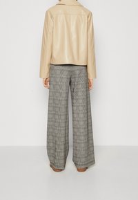 Persoon in hooggeplaatste grijze geruite wijde broek, beige leren jas en bruine schoenen, staande tegen een effen lichte achtergrond.