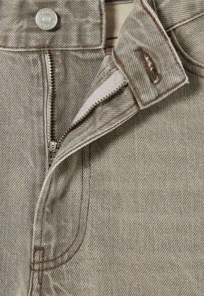 Tela de mezclilla beige con un sutil patrón de espiga. Presenta un cierre de cremallera de metal plateado y un broche a presión. Costuras en contraste visibles.