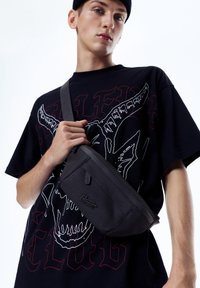 Crni torbica na rame od teksturiranog materijala, s džepom na zatvarač i logom, nošena preko tijela uz grafičku majicu.