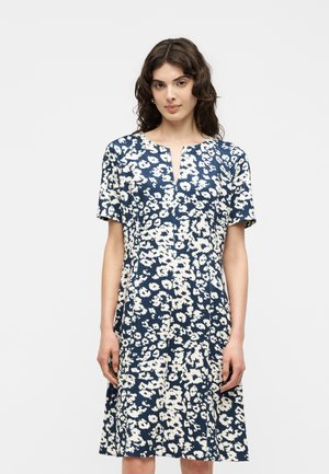 Frau mit schulterlangem welligem Haar, die ein marineblau-weißes, knielanges Kleid mit kurzen Ärmeln und einem geteilten Ausschnitt trägt.
