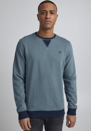 Blauwe crewneck sweatshirt met lange mouwen, contrasterende marineblauwe geribde manchetten en zoom, gestikte details en een klein logo op de linkerkant van de borst.