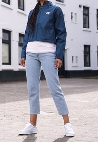 Blauwe waterdichte jas met capuchon, wit T-shirt eronder, lichtblauwe cropped jeans en witte sneakers. Stedelijke buitenomgeving.