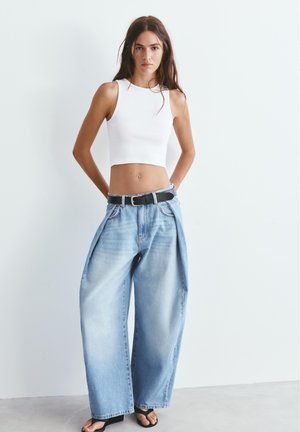 Nő fehér rövid felsőben és széles szárú világoskék farmernadrágban, fekete övvel, egyszínű fehér háttér előtt áll.