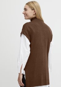 Brun strikket vest med høj krave og korte ærmer, lagdelt over en hvid skjorte med splitærmer. Glat struktur, enkelt design.
