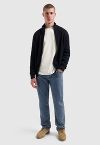 Marineblauwe fleece-jas met ritssluiting over een wit T-shirt, gecombineerd met lichtblauwe jeans en tan kleurige moccasin-stijl schoenen met een geribbeld textuur.