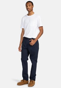 White short-sleeve t-shirt with a chest pocket, paired with navy blue trousers and tan suede shoes, standing against a light background.