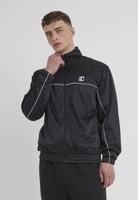Black athletic jacket made of smooth synthetic material. Features white piping across the chest and elastic cuffs and waistband.