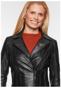 Schwarze Lederjacke mit taillierter Passform, ausgestattet mit Reißverschlüssen und geflochtenen Akzenten, lässig über einem rostfarbenen Rippstrick-Pullover getragen.
