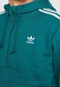 Tyrkysová mikina Adidas se zipem u krku, zdobená bílými pruhy na ramenou a bílým logem uprostřed hrudi. Jemná textura.