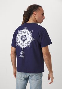 Navy blue cotton t-shirt with a white graphic on the back, featuring a circular emblem with floral and text details. Short sleeves, relaxed fit.