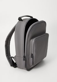 Mochila gris con un acabado mate y suave. Presenta un compartimento principal y un bolsillo delantero con cremallera. Tiene correas acolchadas y un asa en la parte superior para llevarla.