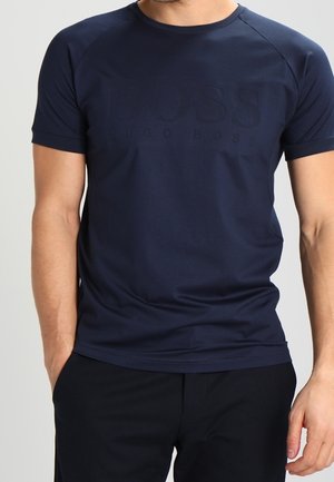 Homme portant un T-shirt bleu marine ajusté à manches courtes avec un logo en relief discret "BOSS Hugo Boss" sur le devant et un pantalon noir.
