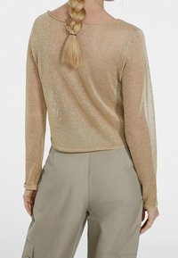 Gul, långärmad topp med en genomskinlig, glittrig textur. Croppad design med rund halsringning och rak fåll. Bärs tillsammans med beige byxor.