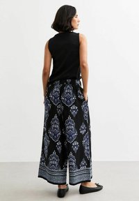 Schwarzes ärmelloses Top kombiniert mit weit geschnittenen Hosen, die ein blaues und weißes dekoratives Muster sowie einen kontrastierenden Saum aufweisen. Flache schwarze Schuhe.