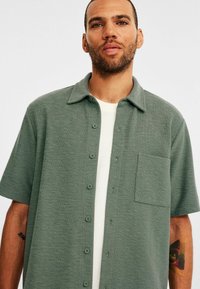 Korte mouwen shirt in gedempt groen, vervaardigd uit textuurstof, met een knoopsluiting aan de voorkant en een borstzak. Glad kraag en subtiele details.