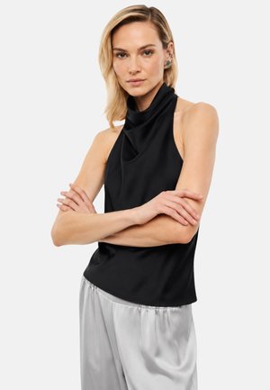 Vrouw met lichtblond haar die een mouwloze zwarte top met hoge hals en losse lichtgrijze broek draagt, armen gekruist voor een witte achtergrond.