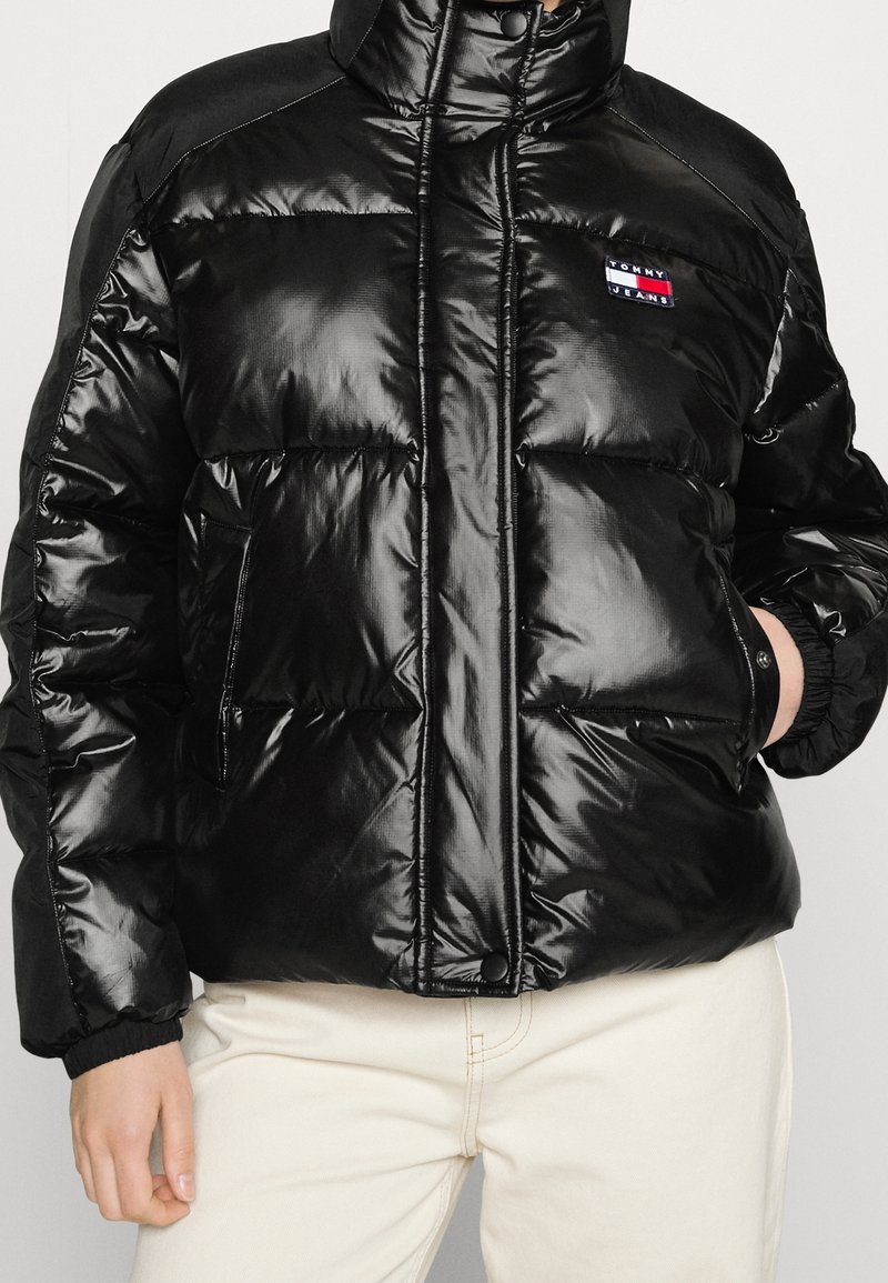 Veste noire matelassée avec col haut, texture brillante, poches zippées et logo brodé sur la poitrine. Dotée de poignets et d'un ourlet élastiques.