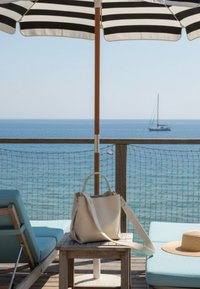 Μπεζ τσάντα πάνω σε ξύλινο τραπέζι ανάμεσα σε δύο μπλε αναπαυτικές καρέκλες κάτω από μαύρο-λευκή ριγωτή ομπρέλα κοντά στον ωκεανό με ιστιοφόρο στο φόντο.