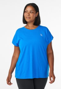 Frau mit schulterlangem Haar, die ein leuchtend blaues Kurzarm-T-Shirt und schwarze Hosen trägt und leicht nach links schaut.