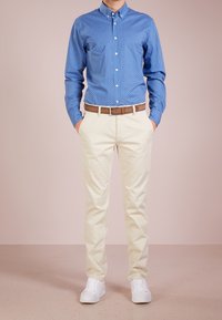 Blå skjorta med knappar och små vita prickar, krämfärgade chinos och vita sneakers. Brunt bälte accentuerar midjan. Enfärgad bakgrund.