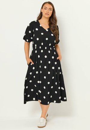 Kvinde iført en sort midi-kjole med hvide prikker, korte puff-ærmer, lommer og hvide sneakers, stående mod en ensfarvet baggrund.