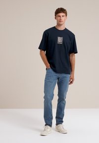 Mornarsko modra bombažna majica s kratkimi rokavi, z kvadratnim logotipom na sredini. Oblečena z svetlo modrimi kavbojkami in belimi supergami.