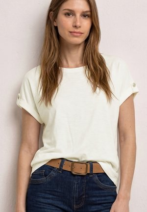 Jeune femme aux longs cheveux bruns portant un haut blanc à manches courtes rentré dans un jean bleu foncé avec une ceinture beige.