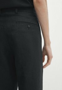 Close-up van zwarte linnen broek met een achterzak met knoop en een hand op de heup, waarbij de textuur en pasvorm van de stof worden benadrukt.