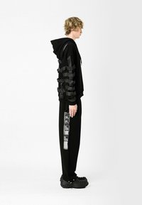 Sudadera negra con acentos en capas similares a cintas en las mangas, combinada con pantalones negros que exhiben detalles gráficos; el calzado es negro texturizado.