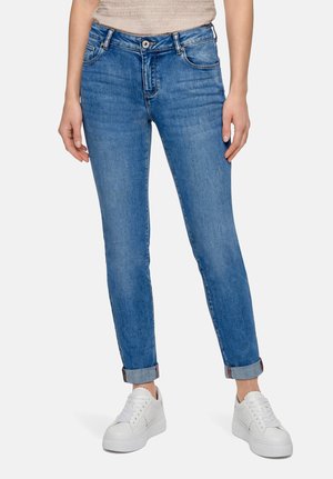 Femme portant un jean bleu avec revers et des baskets blanches, debout devant un fond clair uni.