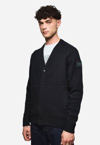 Schwarze Strickjacke mit V-Ausschnitt, Front mit sechs Knöpfen und zwei Seitentaschen. Mit gerippten Bündchen und einem Patch auf dem linken Ärmel.