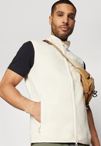 Flisasti brezrokavnik v off-white barvi z visokim ovratnikom in zadrgo. Ima stranske žepe in je oblečen preko črne srajce.