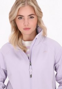 Svetlo vijoličasta zip-up jakna z visokim ovratnikom, ki vsebuje majhen logotip dežnika in vrvico pri zadrgi. Pod njo je bela in vijoličasta črtasta majica.