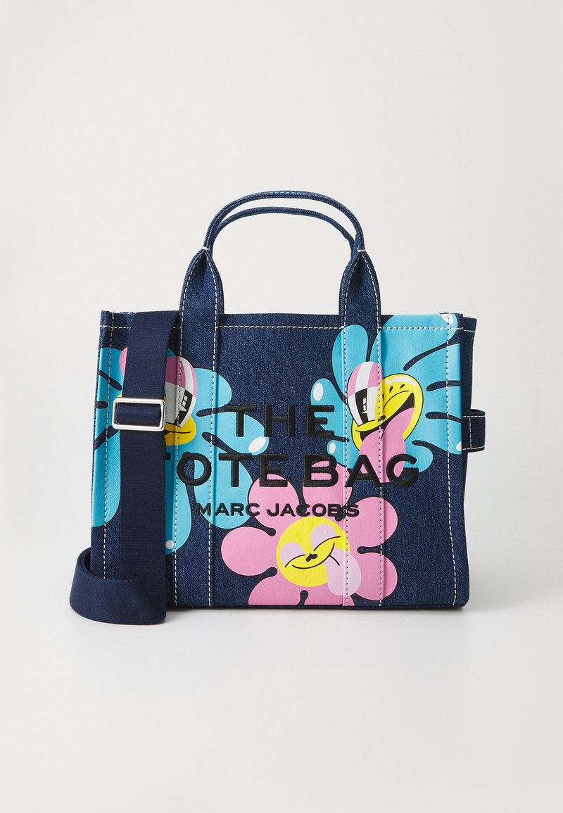 Sac en denim avec des imprimés de fleurs souriantes colorées, texte noir "THE TOTE BAG MARC JACOBS" et une bandoulière ajustable bleu marine.