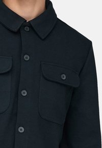 Schwarzes, langärmliges Hemd mit Knopfleiste, spitzem Kragen und zwei aufgesetzten Brusttaschen mit Klappen, die jeweils mit einem einzelnen Knopf verschlossen sind.