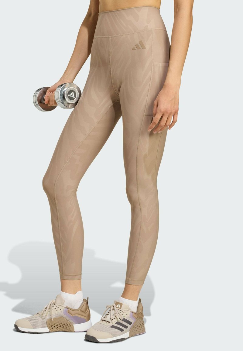 Beige Leggings mit hohem Bund und dezentem Animal-Print, aus elastischem Stoff. Kombiniert mit hellbeigen und violetten Sportschuhen.