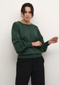 Maglione verde a maglia con un ampio scollo e maniche a sbuffo, finitura testurizzata, abbinato a pantaloni neri, su uno sfondo bianco semplice.