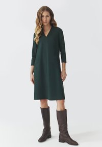 Robe verte, longueur genou avec un col en V et des manches 3/4, associée à des bottes marron jusqu'aux genoux. Design simple et épuré sans motifs visibles.