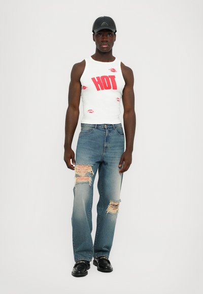 Witte tanktop met rode "HOT" tekst en lippenafdrukken, gecombineerd met versleten lichtblauwe jeans met rafelige patches en zwarte schoenen.