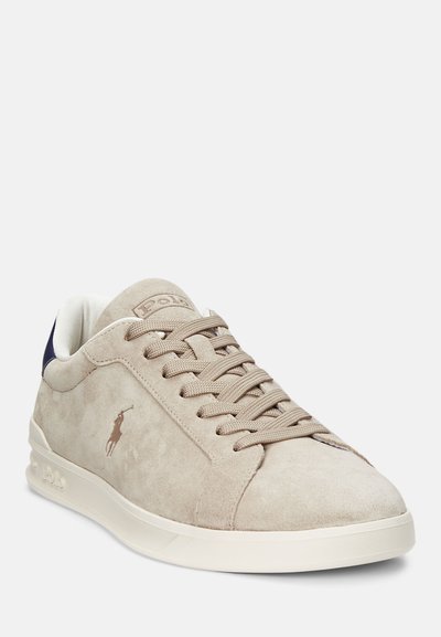 Beige suede sneaker with a white sole and navy accent at the back. Features textured laces and a subtle logo on the side. Rounded toe design.
