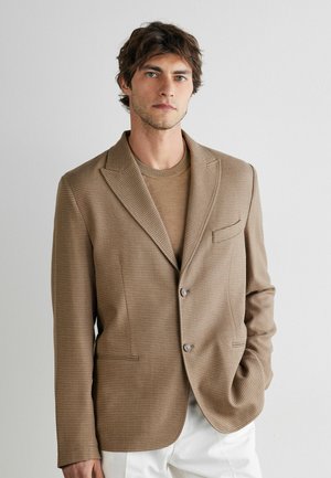 Ung mand med brun tekstureret blazer og matchende sweater, hvide bukser, stående mod en ensfarvet lys baggrund, hånd i lommen.