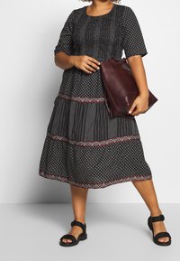 Vestido negro con intrincados patrones blancos, mangas cortas y ribetes multicolores. Combinado con sandalias negras y un bolso burdeos.