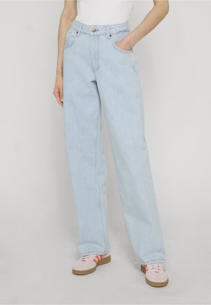 Person som har på seg lyseblå vid jeans, hvit topp og rosa joggesko med oransje striper, står med en hånd i lomma.