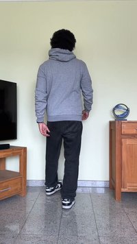 Sweatshirt cinza com capuz e corte descontraído, jeans pretos e sneakers em preto e branco. O fundo inclui uma TV e móveis de madeira.