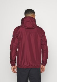 Maroon windbreaker jacket with a hood, elastic cuffs and hem, smooth texture, and logo patch on the left shoulder.