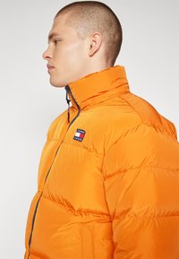 Giacca a vento arancione con collo alto, chiusura con zip e patch con logo. Tessuto liscio con sezioni trapuntate orizzontali.