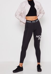 Mustat legginsit, joissa on valkoisella painettuna "THE NORTH FACE", korkea vyötärö, yhdistettynä vaaleanpunaisiin toppaustuulihaalariin ja mustiin lenkkareihin.