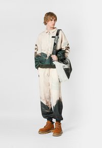 Sudadera de gran tamaño en beige y verde con cuello con cremallera, combinada con pantalones jogger a juego con estampado de paisaje. Botas marrones con cordones y un bolso.